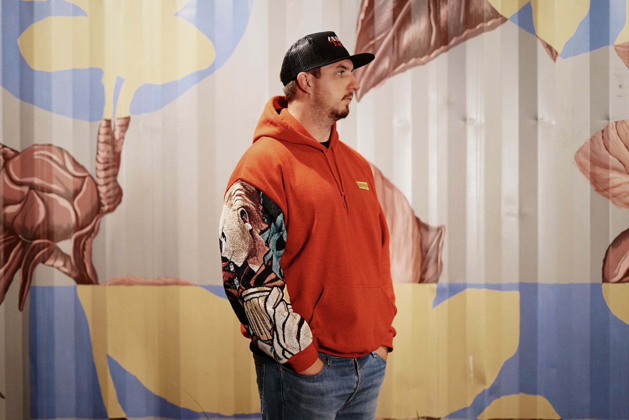 Male model wearing Million Tapestry Sleeve Hoodie in front of a flower painted wall